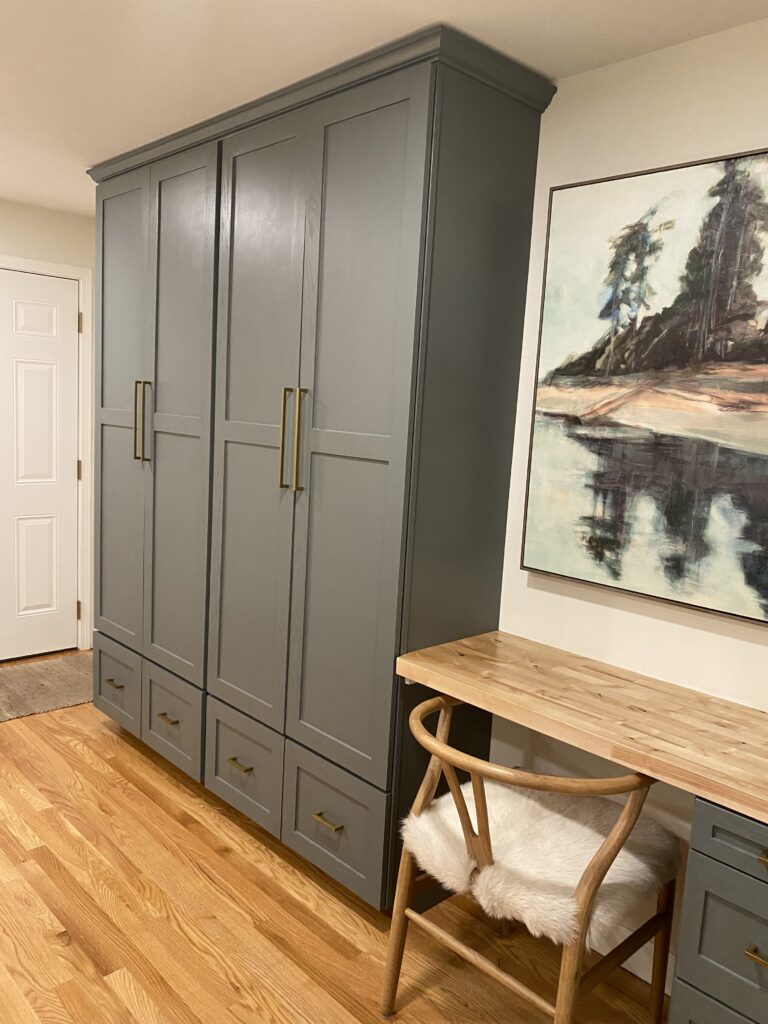 Elegant built-in cabinetry in a muted sage green tone with modern brass hardware. The unit features double-door upper cabinets and sits beside a painted landscape artwork and a nearby white door.