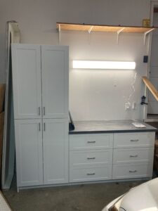 Simple and functional garage storage setup with a mounted wooden shelf supported by white brackets and an under-cabinet light. Also includes two painted cabinet doors leaning against the wall, ready for installation.