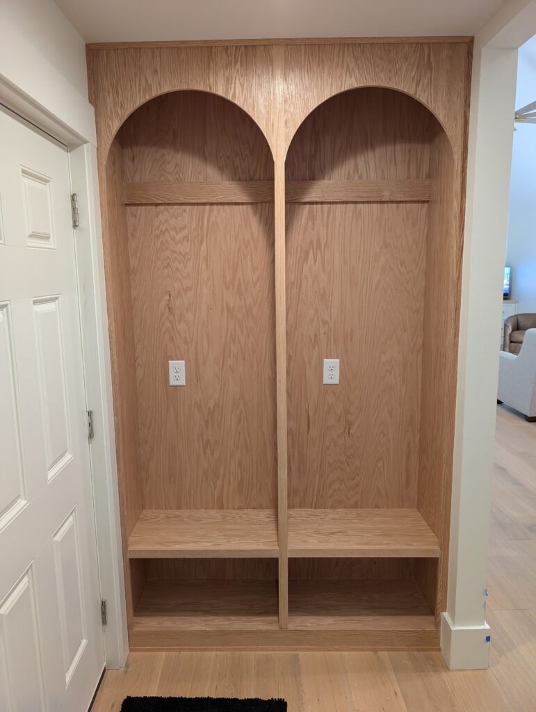 Custom mudroom bench and storage unit crafted from light wood. Features two large cubbies with electrical outlets, open bench seating, and upper archways, built into a recessed wall next to a white door.