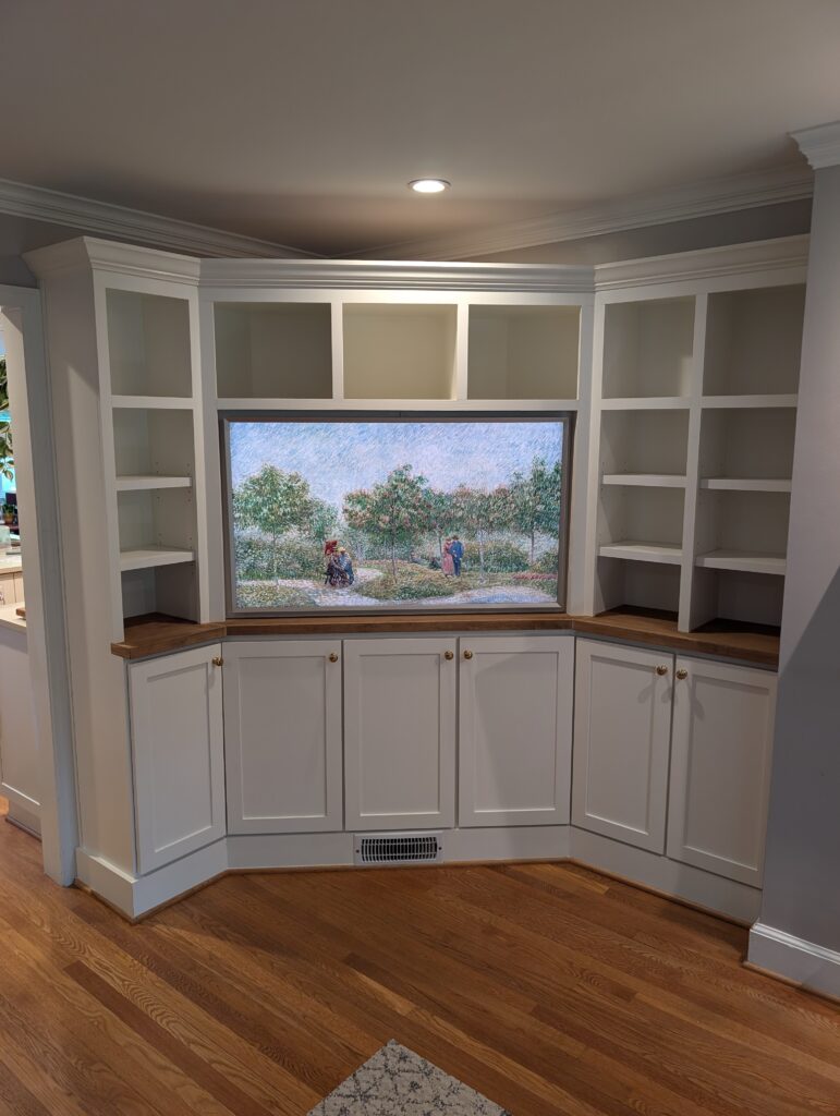 White built-in wall unit with open shelving and lower cabinets, designed to fit the corner of a living space. A framed artwork is set into the center section above a wooden countertop, with hardwood floors completing the look.